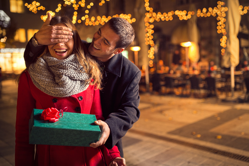 A man surprising a smiling woman with a present as he covers her eyes